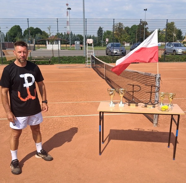Mężczyzna stojący przy stole z pucharami i biało-czerwoną flagą na korcie tenisowym.