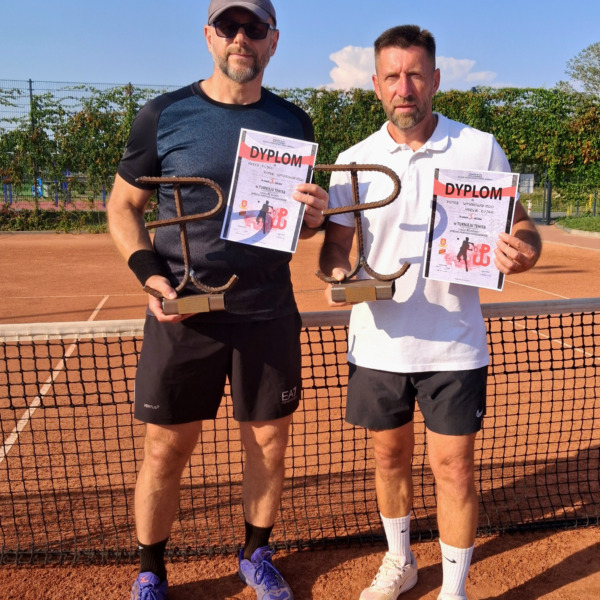 Dwóch uczestników turnieju z dyplomami i pamiątkowymi trofeami na korcie tenisowym.